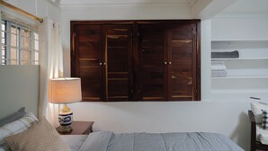 A bedroom with a bed, a nightstand with a lamp, a wooden wardrobe, and a shelf with folded towels. - Sundrift Apartments (Gros Islet)