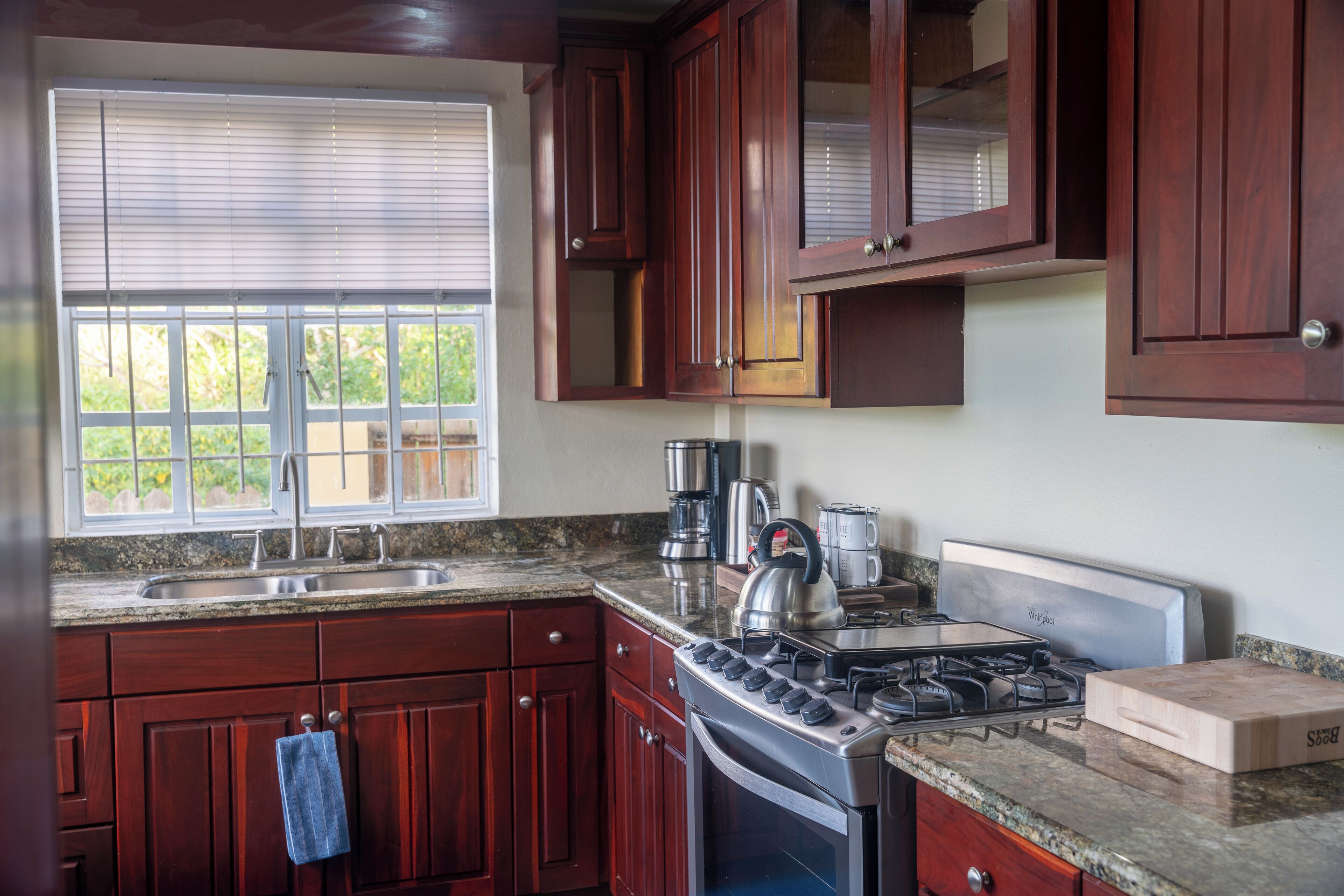 A kitchen with wooden cabinets, a stainless steel sink, a stove, and a window with blinds.
