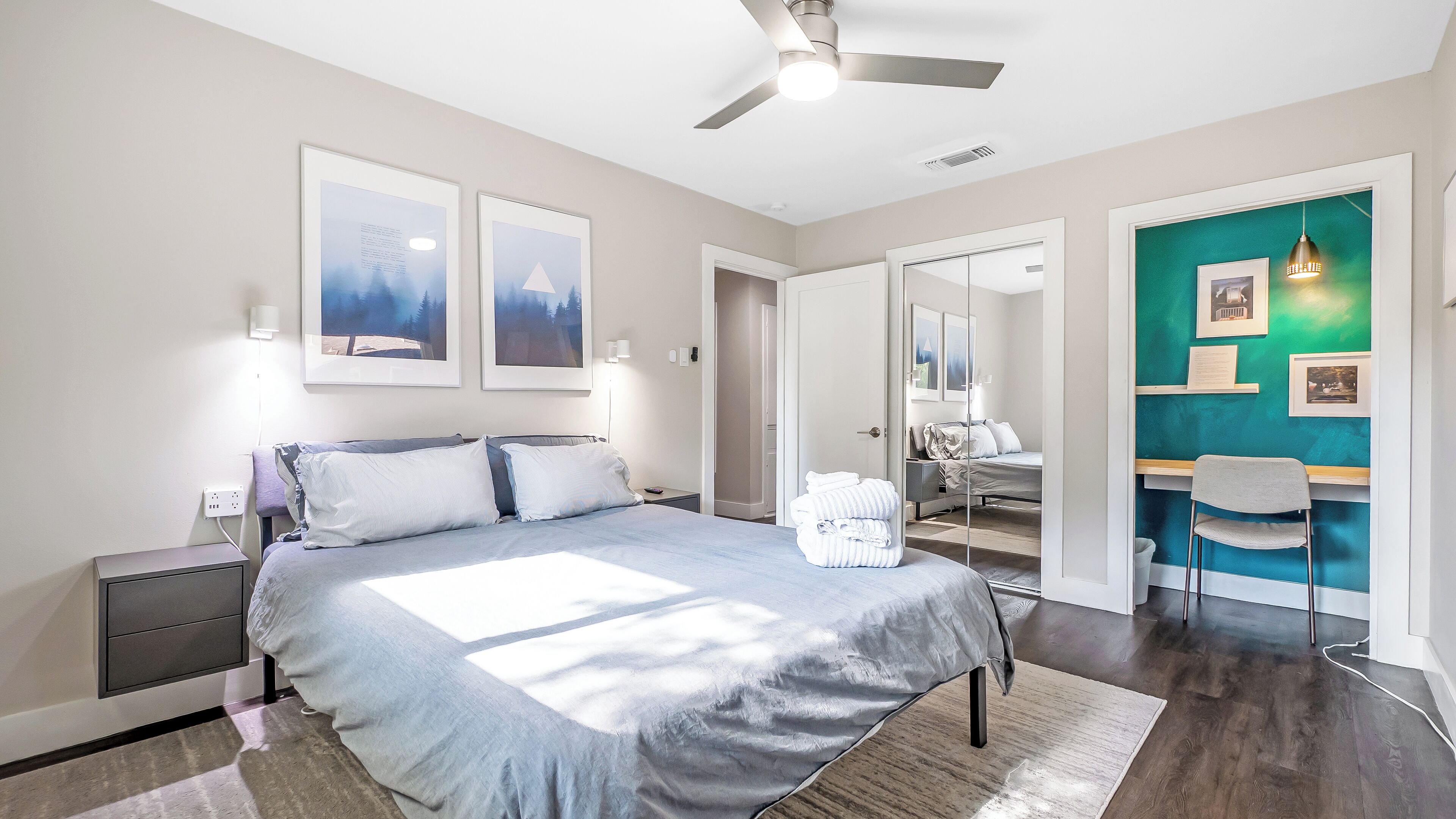 Une chambre à coucher moderne avec un lit, des tables de chevet, un bureau et une chaise. On y trouve des tableaux encadrés au mur, un ventilateur de plafond et un miroir.