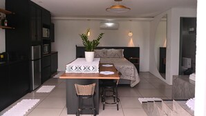 Uma cozinha moderna com armários escuros, uma geladeira e um micro-ondas. Há uma área de jantar com mesa, cadeiras e um vaso de planta. O quarto possui uma cama com cabeceira e uma mesinha de cabeceira.