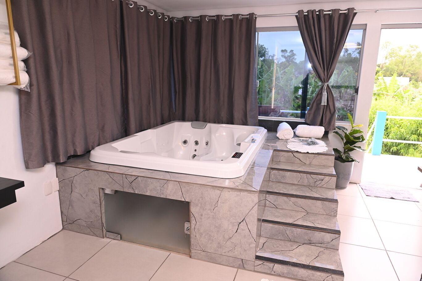 A modern bathroom with a large bathtub, marble flooring, and a view of greenery outside.