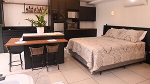 Quarto compacto com cama, pequena cozinha e área de refeições para dois.