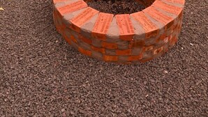 Um poço de fogueira circular de tijolo, com uma moldura de tijolo, rodeado por cascalho e um caminho de tijolo.