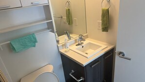 A bathroom with a white toilet, a sink with a mirror above it, and a towel rack with two towels. There are cabinets below the sink and a trash bin next to the toilet. - 5 min walk to university with stunning mountain views (Kelowna)