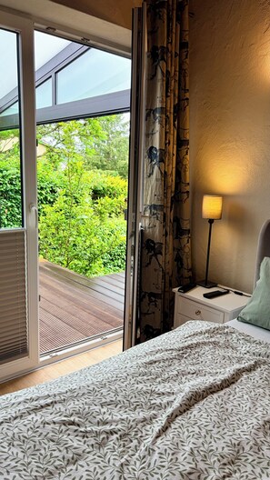 Et soveværelse med en seng, et natbord, en lampe og udsigt til grøn vegetation gennem en skydedør.