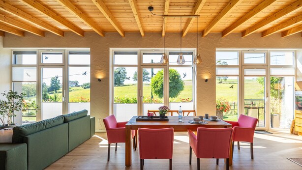 Un comedor con una mesa de madera, sillas rojas y un sofá verde. Amplias ventanas ofrecen vistas a un jardín.