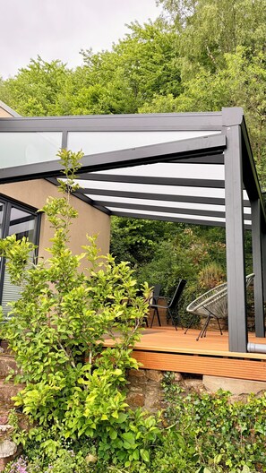 En moderne udendørs terrasse med glasafskærmning, træterrasse og stenvæg.