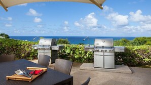 Uma área de refeições ao ar livre com duas churrasqueiras, uma mesa com utensílios e vista para o mar.