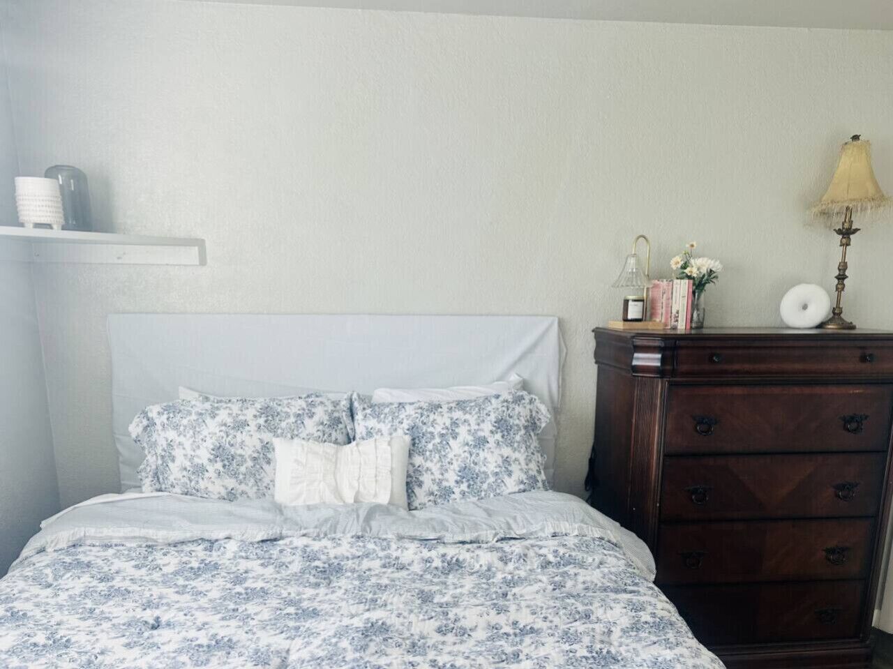 Una camera da letto con un letto, una cassettiera, una lampada e uno scaffale con oggetti.