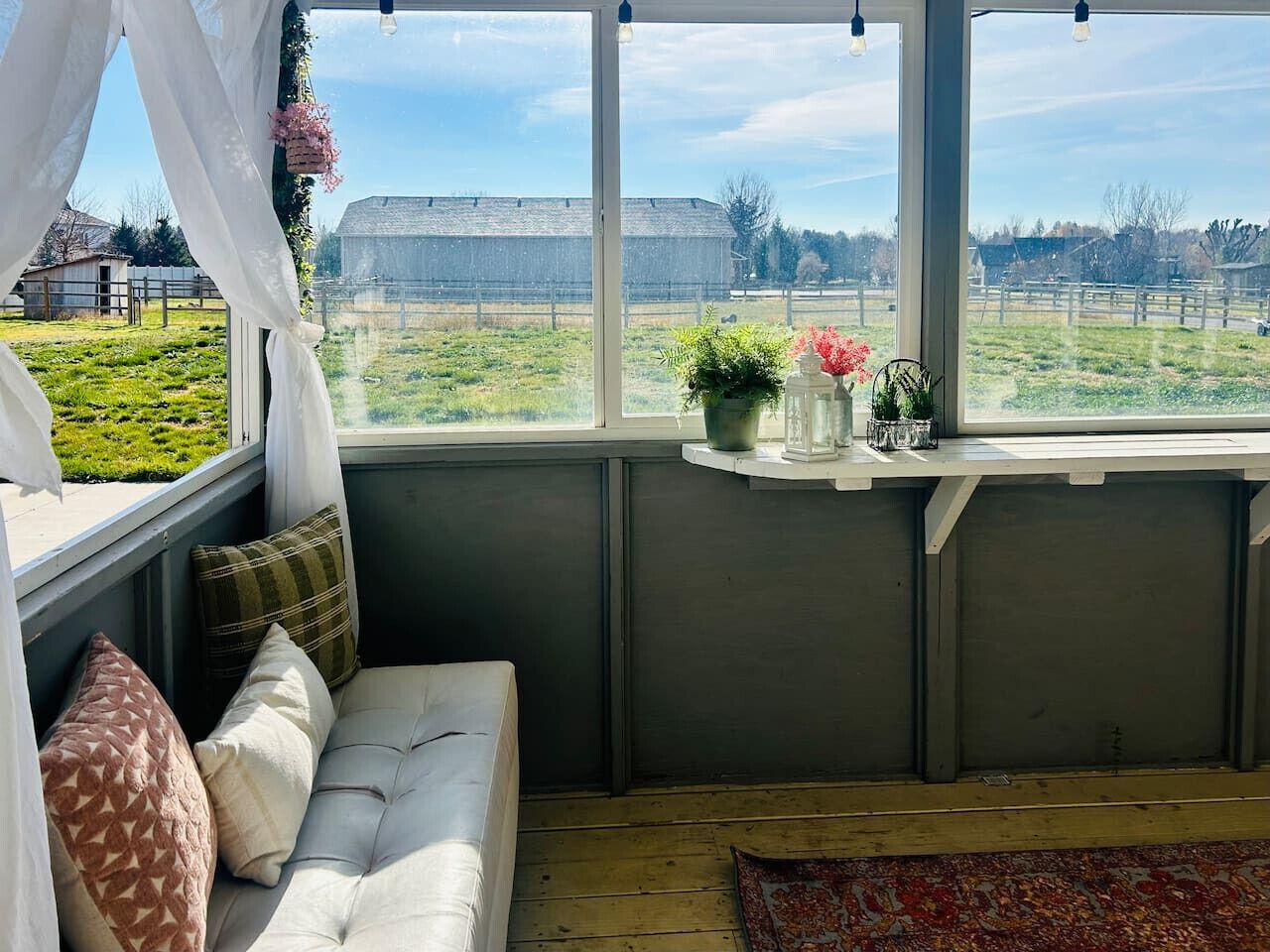 A cozy room with a white sofa, a small window with a view of a green field and a barn, and a shelf with decorative items.