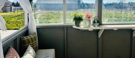A cozy room with a white sofa, a small window with a view of a green field and a barn, and a shelf with decorative items.