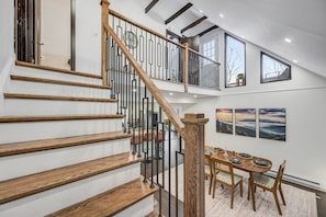 A multi-level interior with a wooden staircase, a dining area with a table and chairs, and large windows.