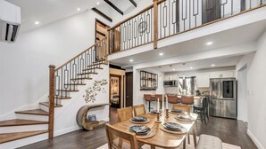 A spacious dining area with a wooden table set for six, a staircase with a modern railing, and a kitchen in the background.