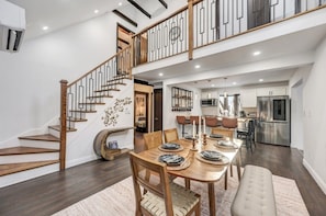 A spacious dining area with a wooden table set for six, a staircase with a modern railing, and a kitchen in the background.