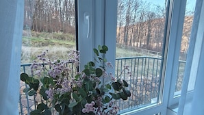Une pièce dotée d’une grande fenêtre, d’un vase blanc orné de fleurs et offrant une vue sur des arbres et une clôture.
