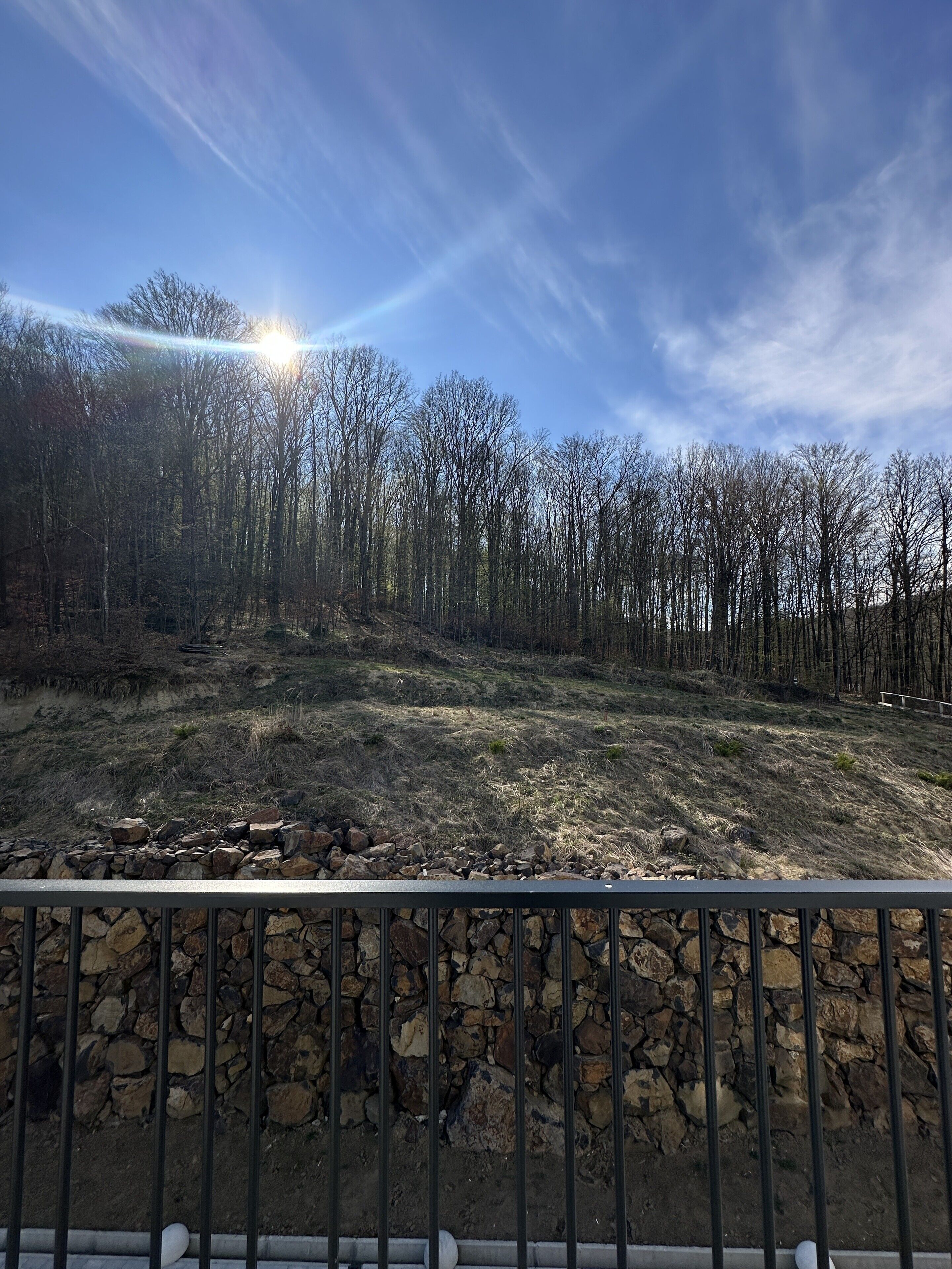 A stone wall with a metal railing, a forested hillside, and a clear sky with the sun shining.