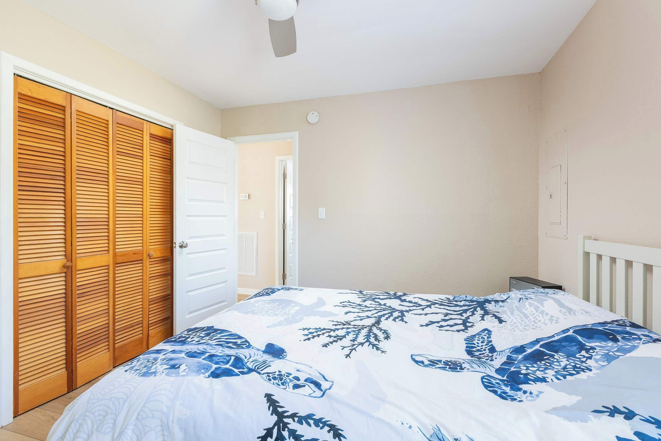 A bedroom with a bed, a wooden wardrobe, a ceiling fan, and a door leading to another room.