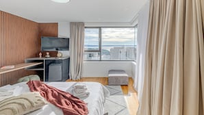 Moderni hotellihuone, jossa on sänky, työpöytä, televisio ja suuri ikkuna, josta avautuu näkymä kaupunkiin.