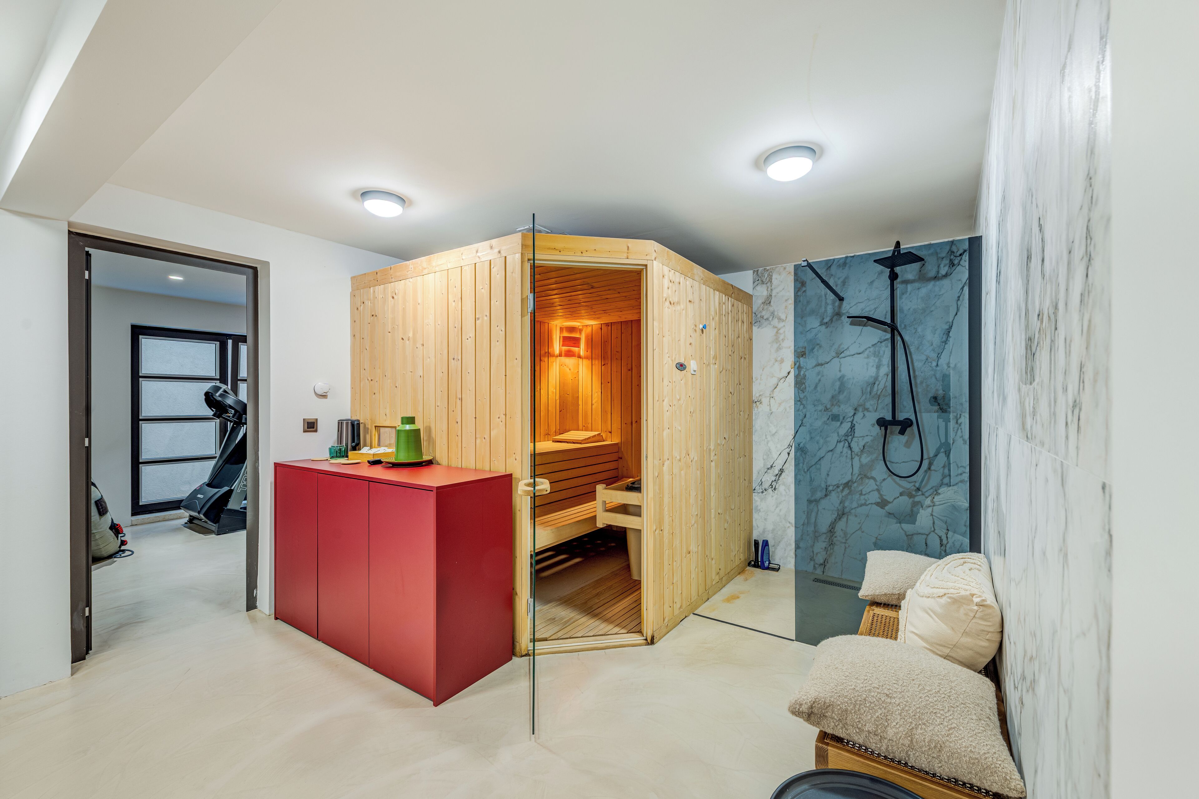 A modern bathroom with a wooden sauna, a red cabinet, a marble shower wall, and a treadmill in the adjacent room.