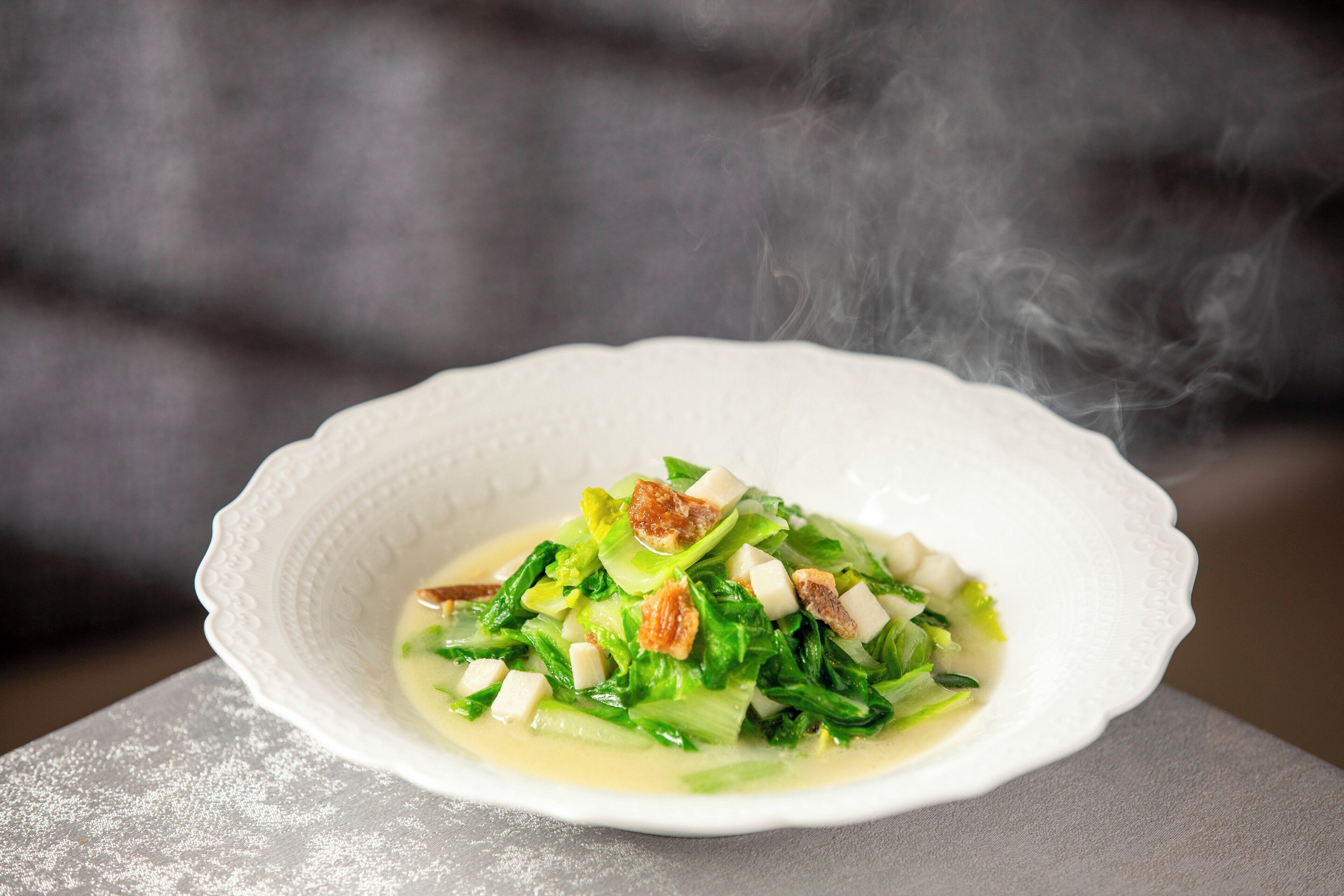 A white bowl with a soup containing green vegetables and croutons, steam rising from the bowl.