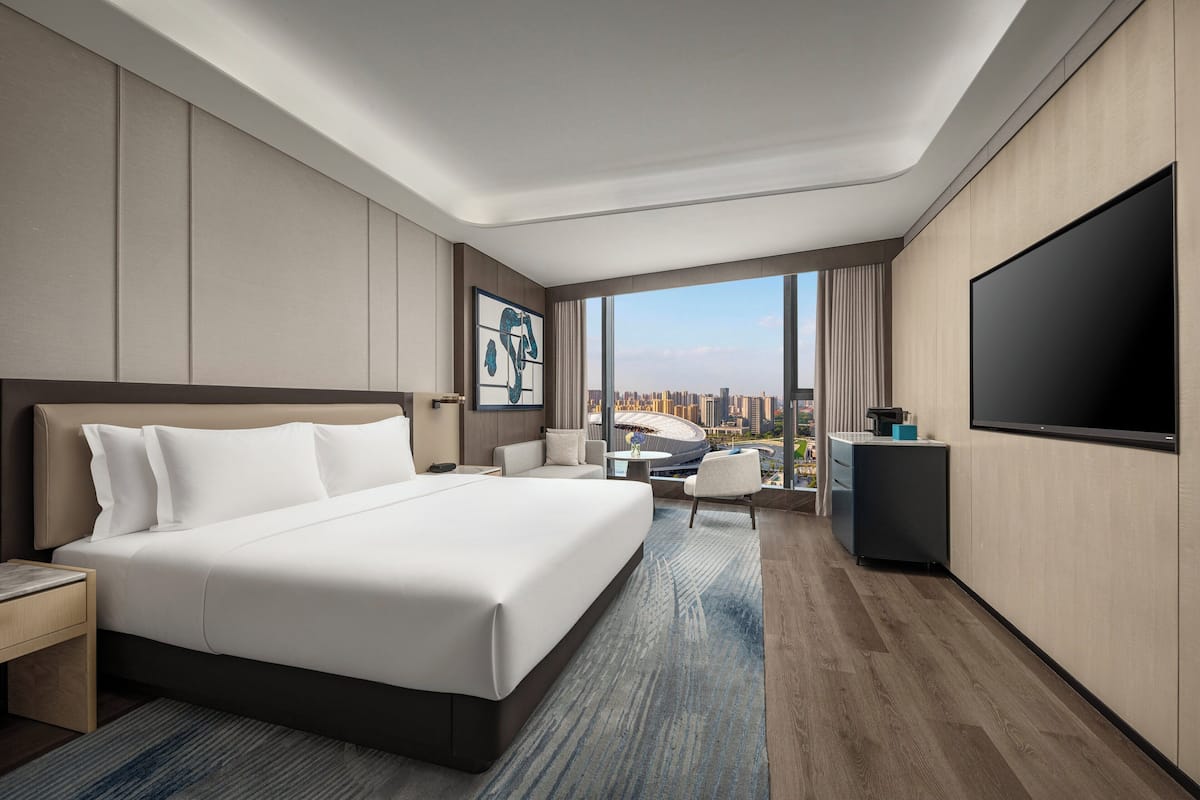 A modern hotel room with a large bed, a flat-screen TV, a desk, and a view of the cityscape.