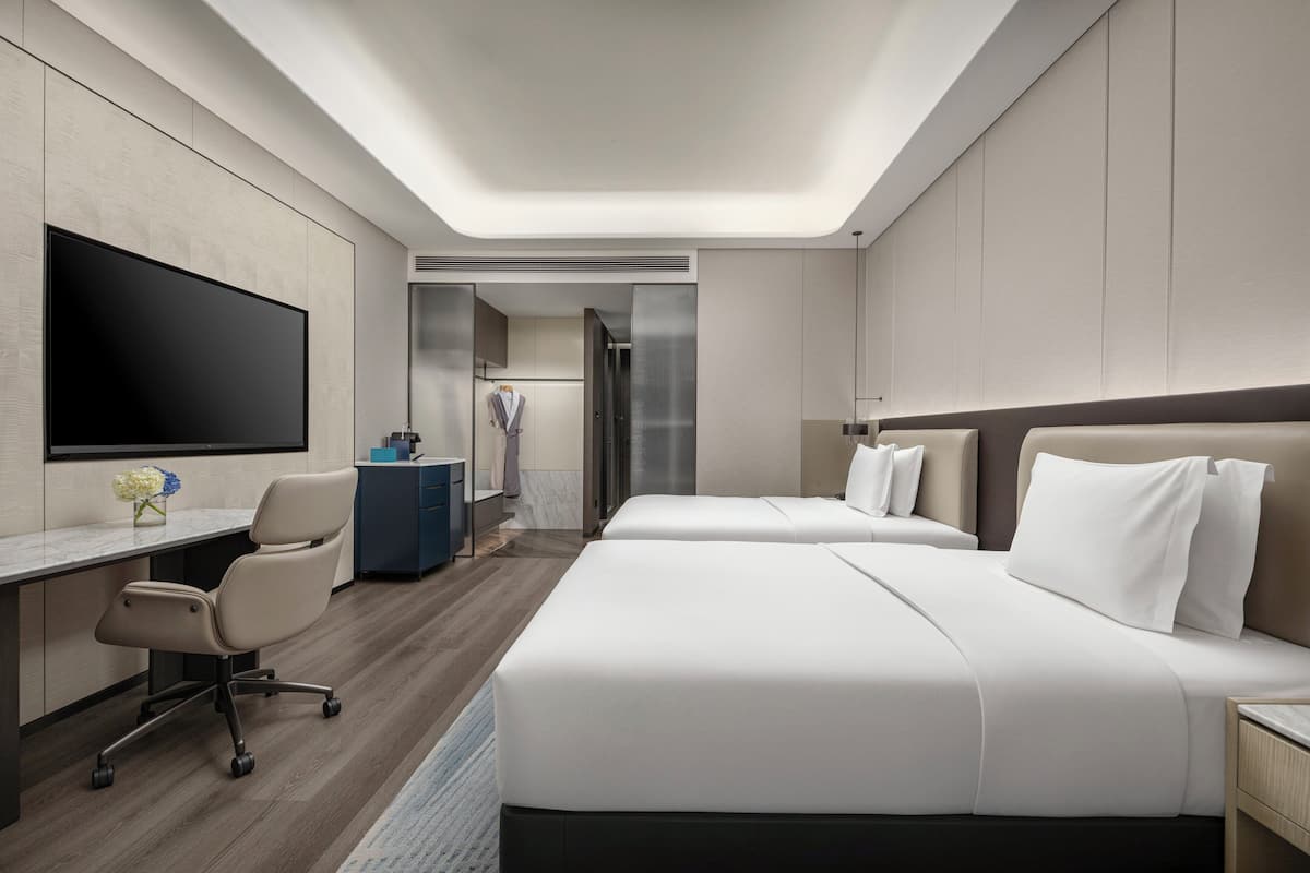 A modern hotel room with a large flat-screen TV, a desk with a chair, two beds, and a walk-in closet.