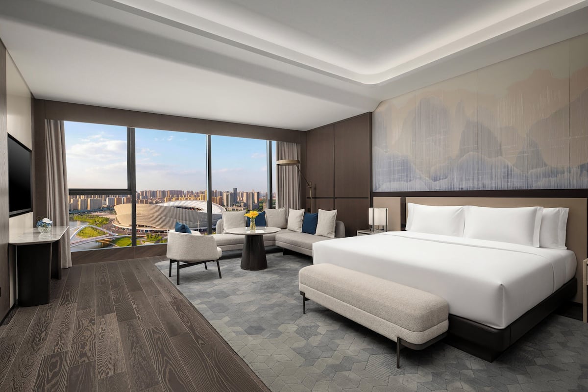 A modern hotel room with a large bed, a sofa, a desk, and a view of the cityscape through large windows.
