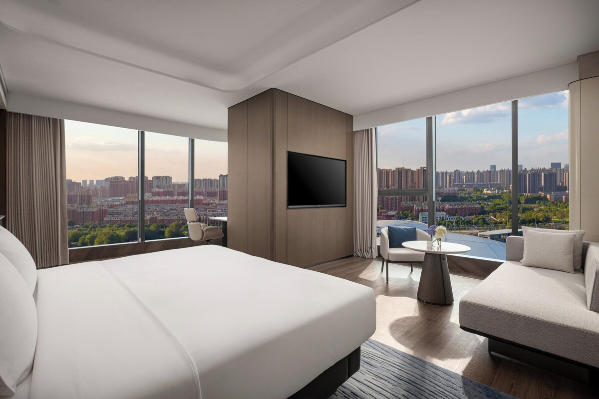 A modern hotel room with a large bed, a flat-screen TV, a sofa, and a dining table.