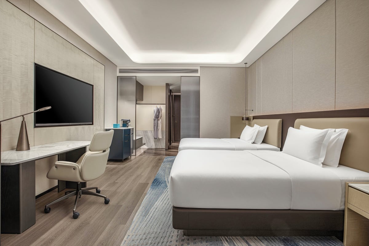 A modern hotel room with a large bed, a desk, a chair, a flat-screen TV, and a walk-in closet.