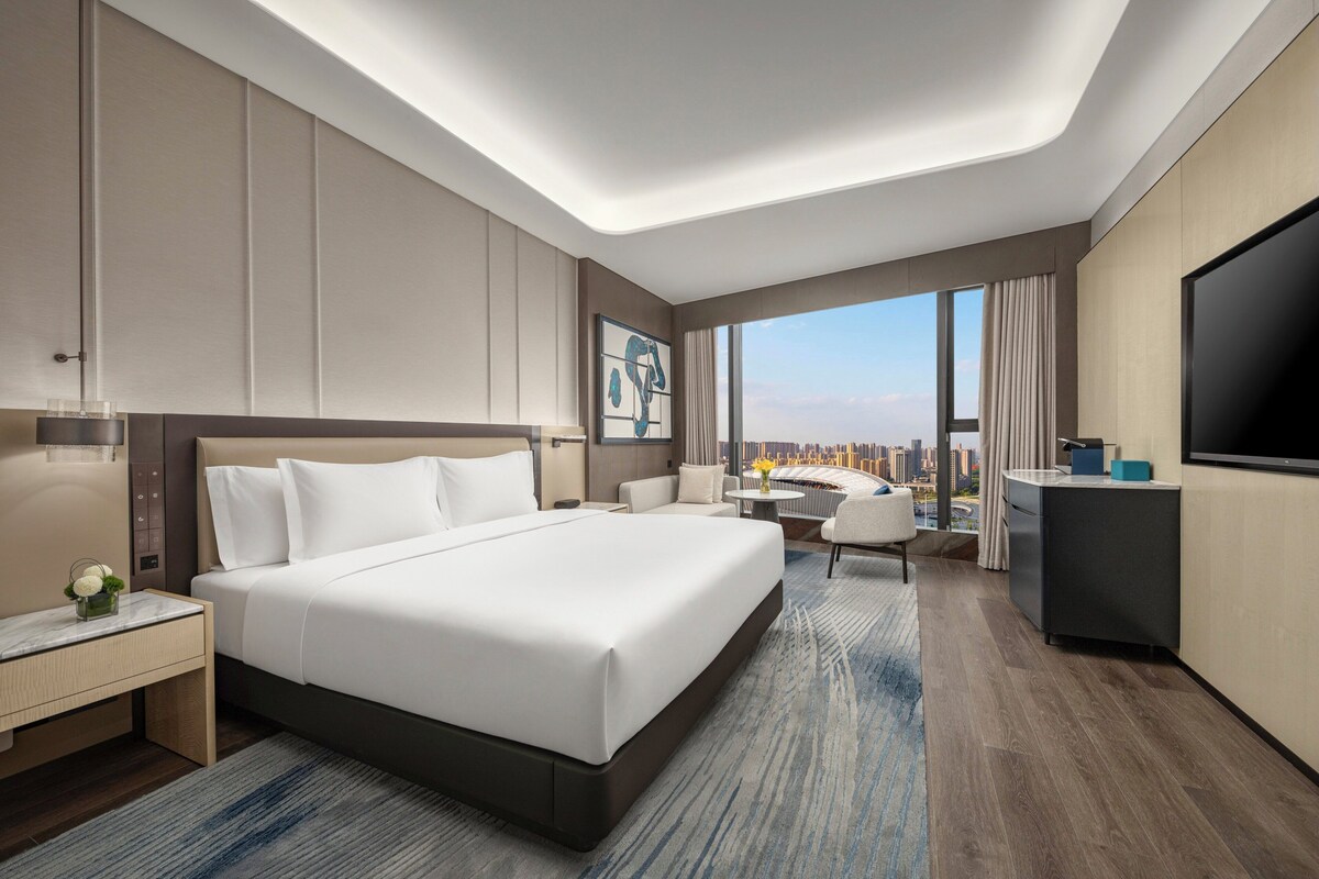 A modern hotel room with a large bed, a desk, a TV, and a city view.