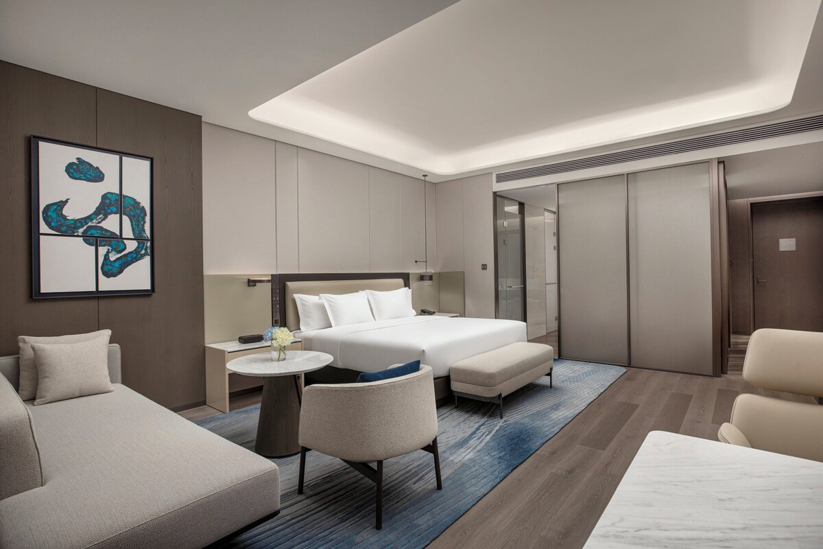 A modern hotel room with a bed, two armchairs, a small table, and a large artwork on the wall.