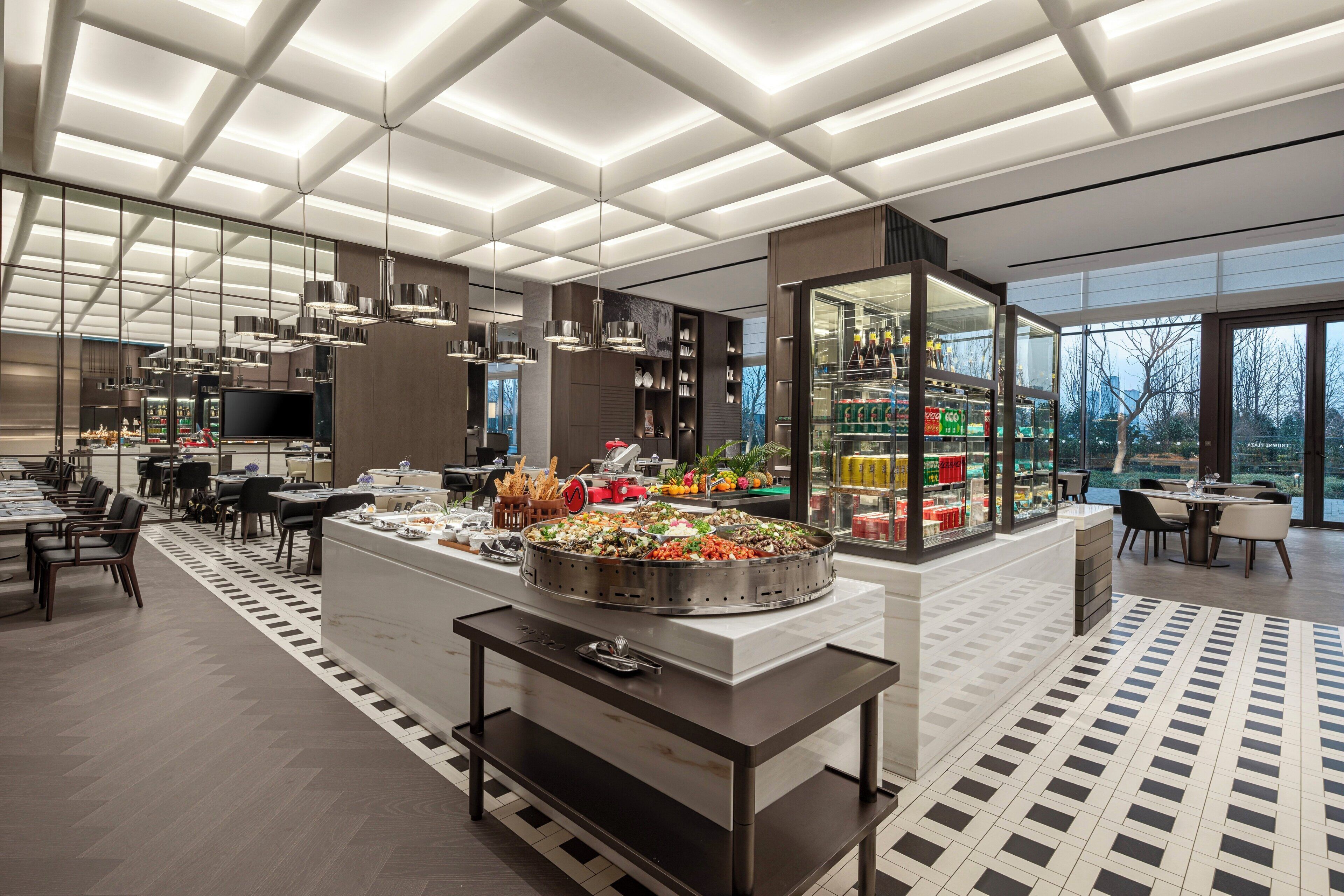 En moderne restaurant med en central buffet, spiseborde og et stort vindue med udsigt til det fri.