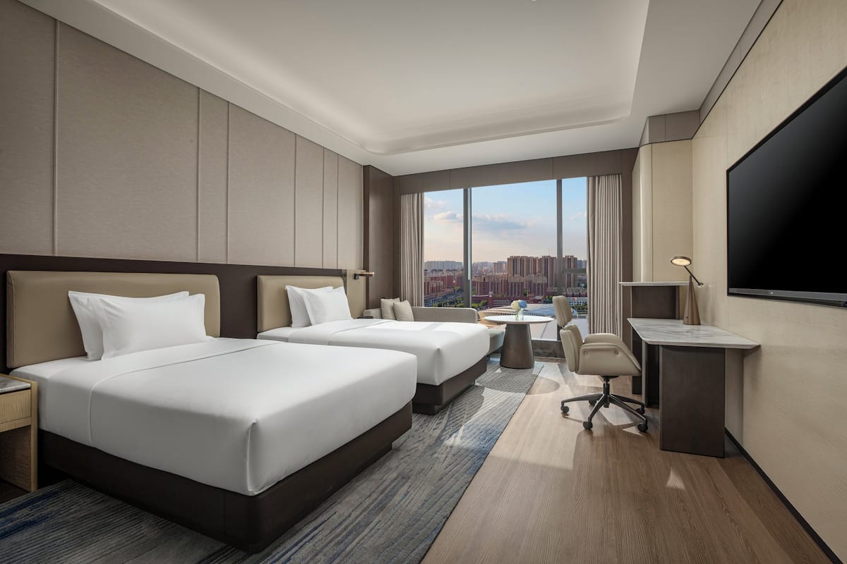 A hotel room with two beds, a desk, a chair, a TV, and a large window with a city view.