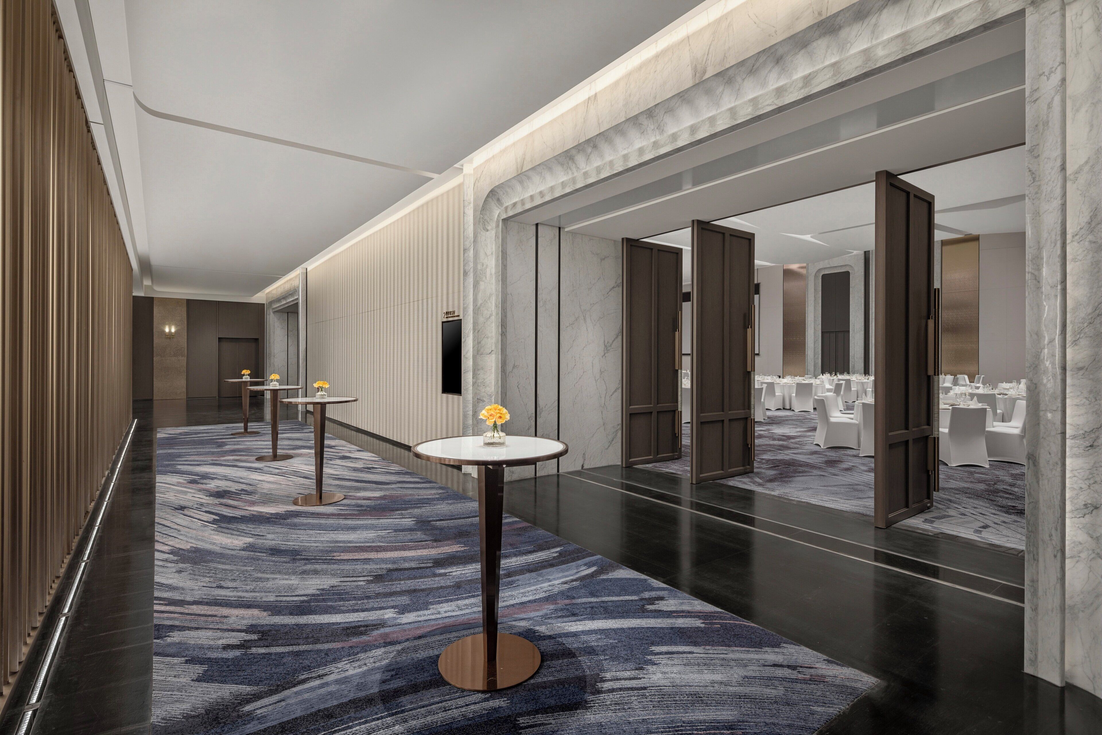 A modern hotel corridor with marble walls, a patterned carpet, and tables with flowers.