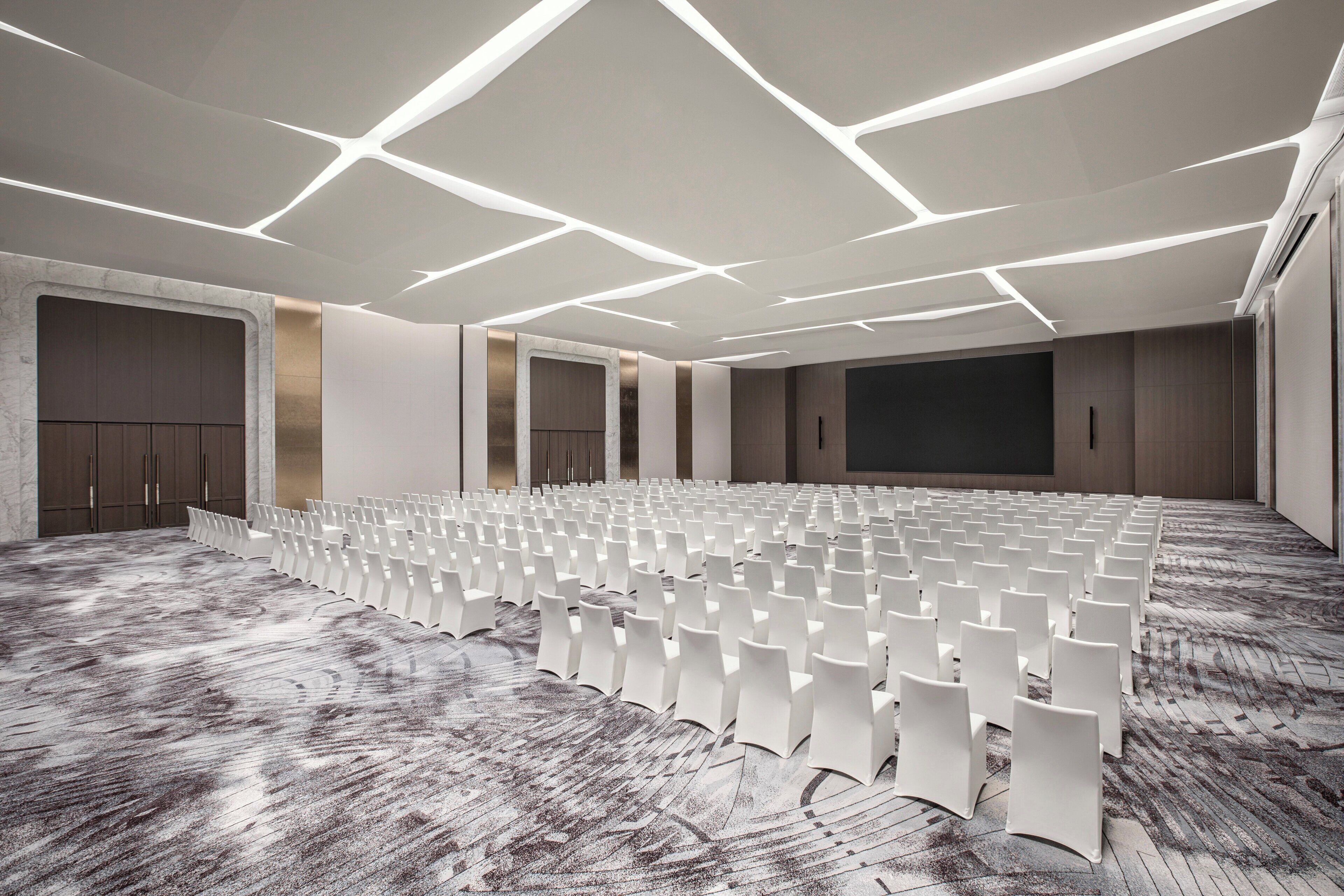 Ein Konferenzraum mit Reihen weißer Stühle, einer großen Leinwand und einem gemusterten Teppich.