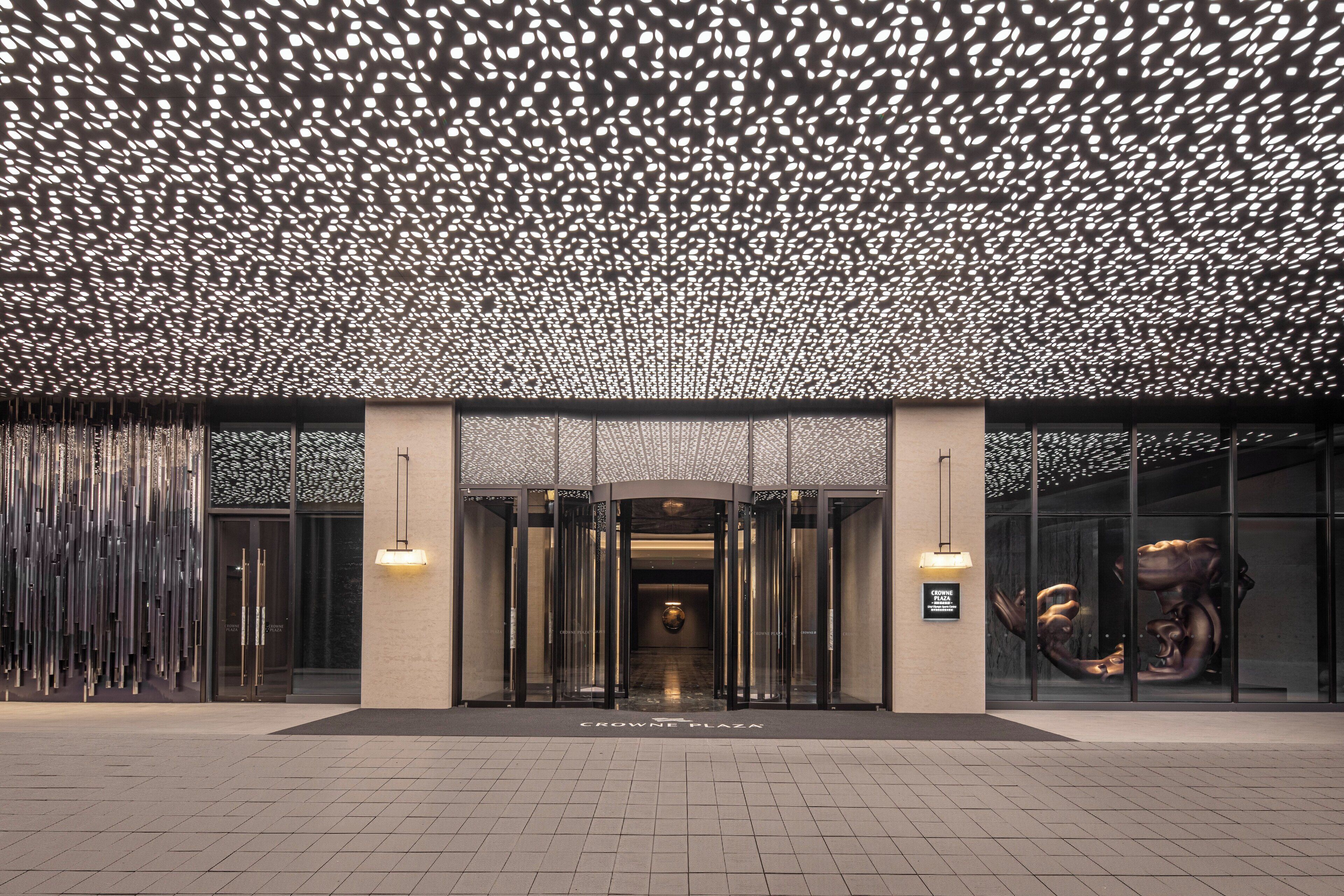 Una entrada moderna a un edificio con techo decorado, fachada de vidrio y una escultura central.