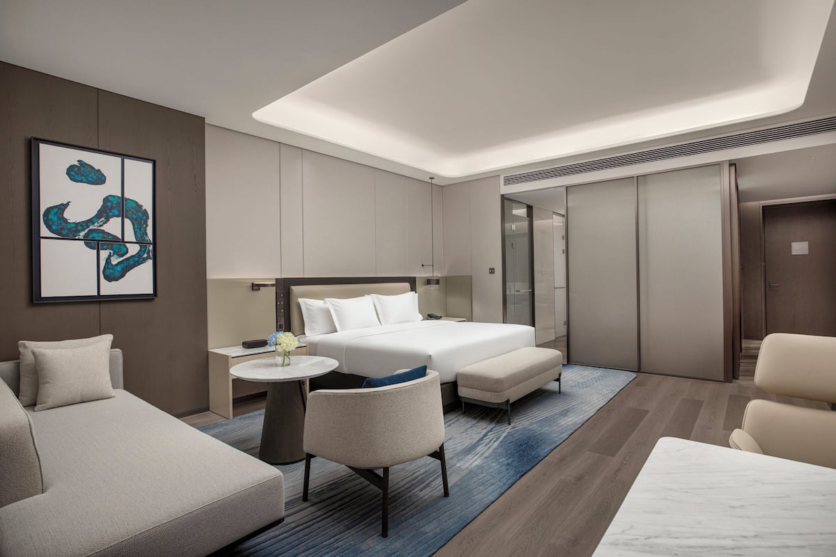 A modern hotel room with a bed, two armchairs, a small table, and a large artwork on the wall.