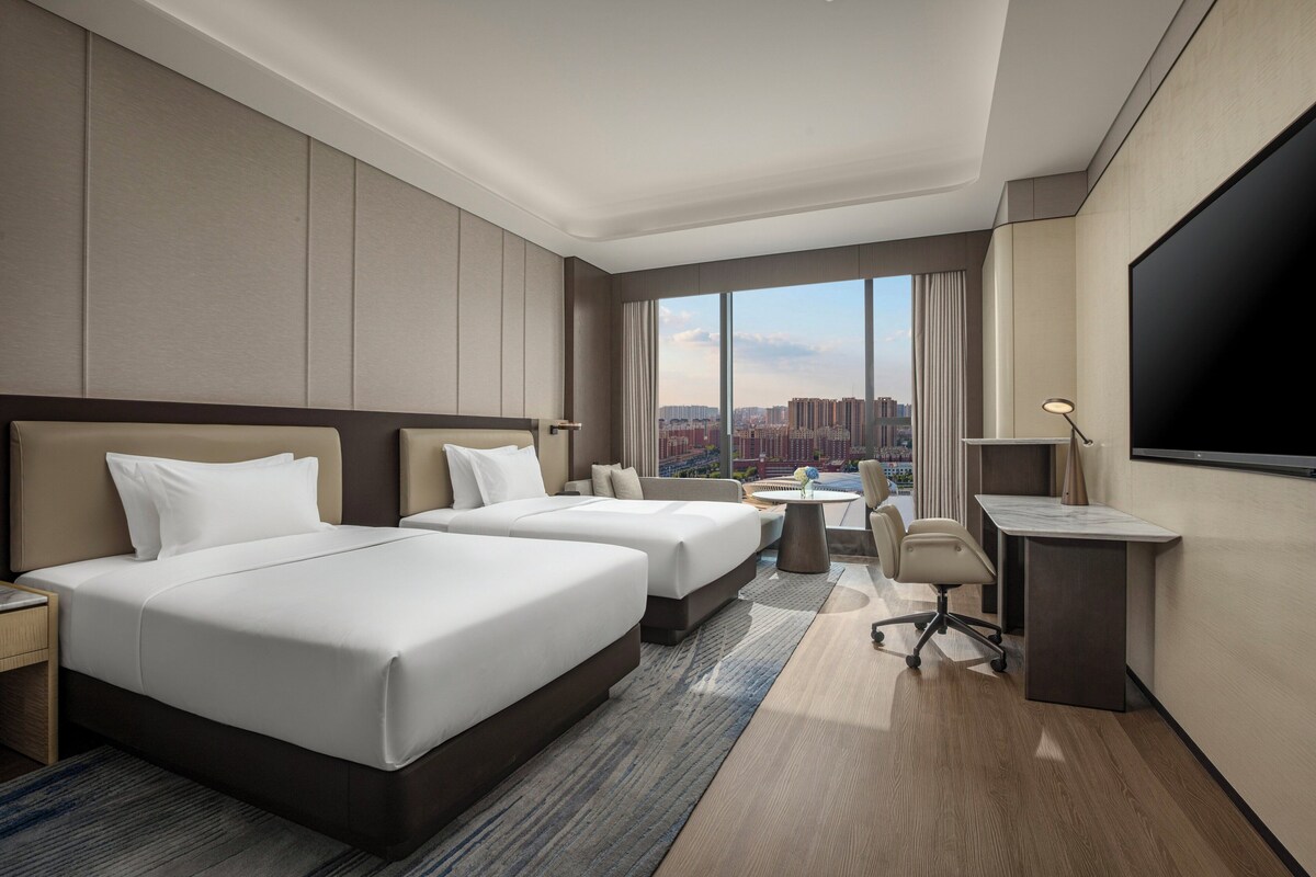 A hotel room with two beds, a desk, a chair, a TV, and a large window with a city view.
