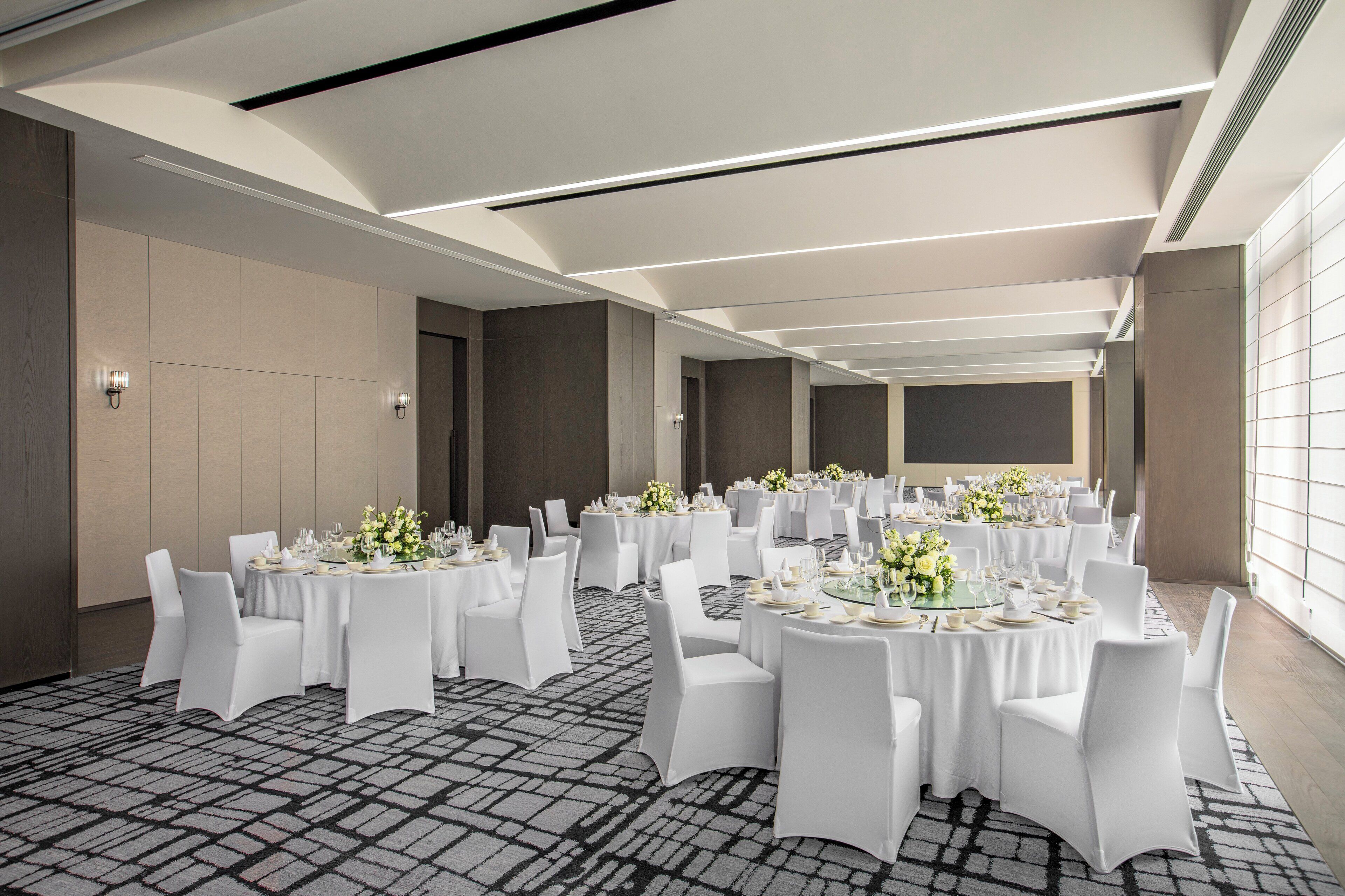 Een ruime feestzaal met ronde tafels gedekt voor een diner, met witte tafelkleden en stoelen.