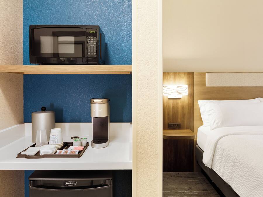 A hotel room with a bed, a nightstand, a wall-mounted lamp, and a kitchenette area with a microwave, coffee maker, and a trash bin.