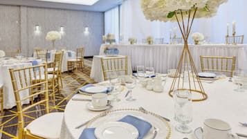 Un salón de banquetes con mesas redondas dispuestas para un evento formal, con sillas doradas, manteles blancos y centros de mesa florales.