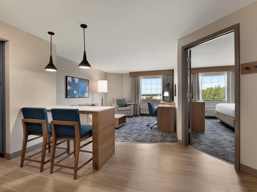 Una habitación de hotel con kitchenette, un comedor con dos sillas, un escritorio con silla, un ventanal grande y una cama.