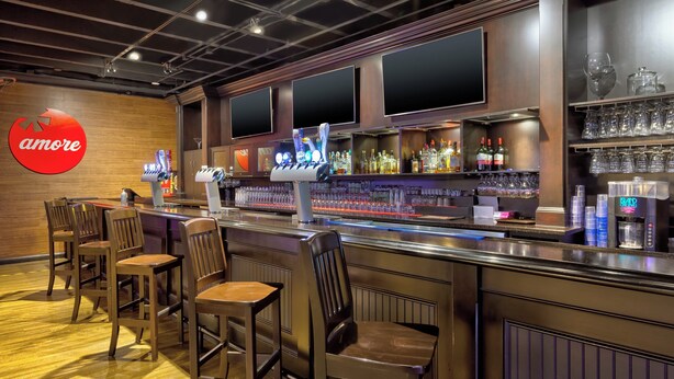 Eine Bar mit einer Theke, mehreren Barhocker und einem Regal mit Gläsern und Flaschen.