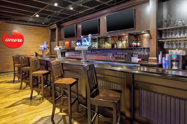 Una zona de bar con mostrador, varios taburetes y estantería con copas y botellas.