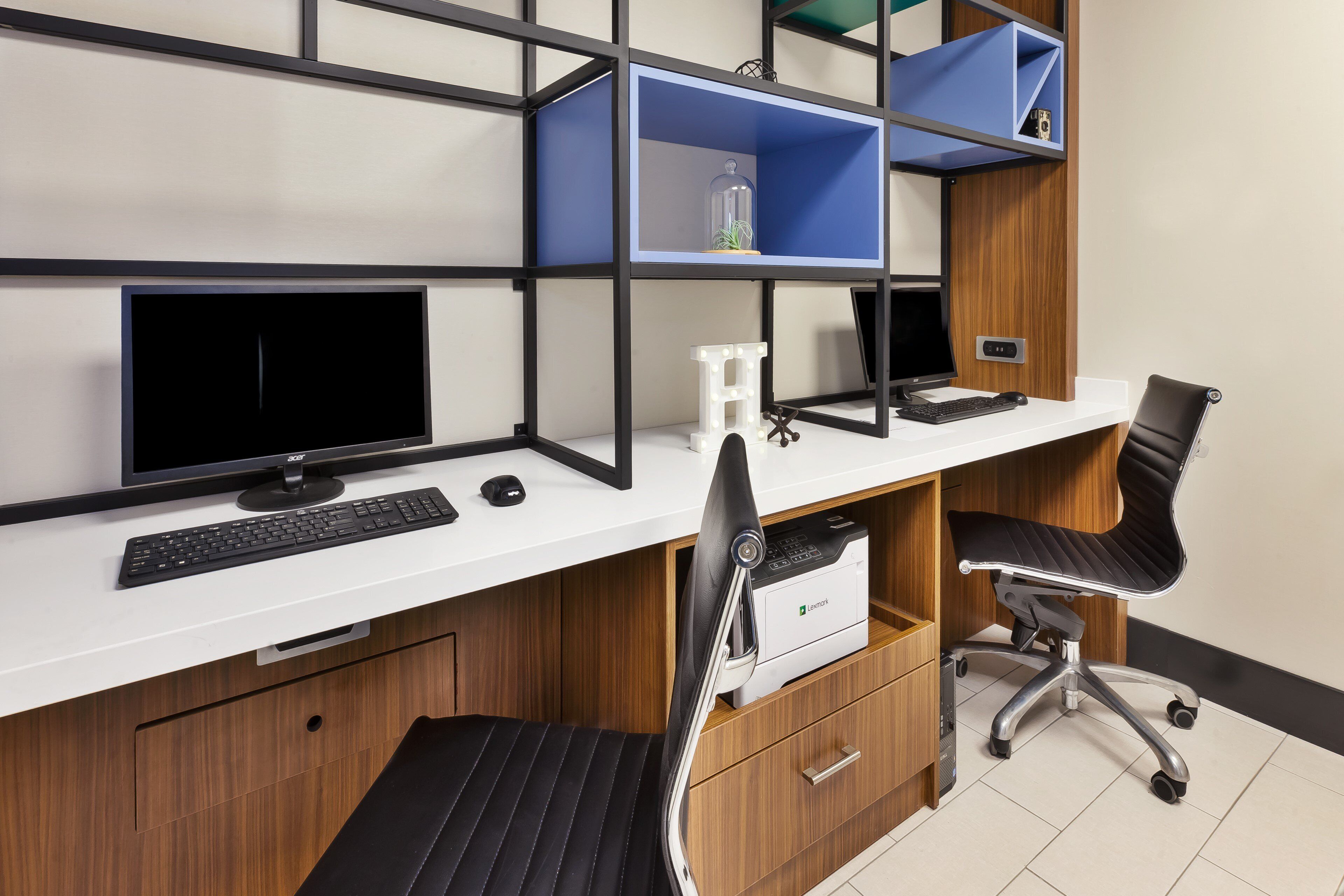 A modern office desk with two monitors, a keyboard, and a chair. There are blue shelves above the desk and a printer on the right.