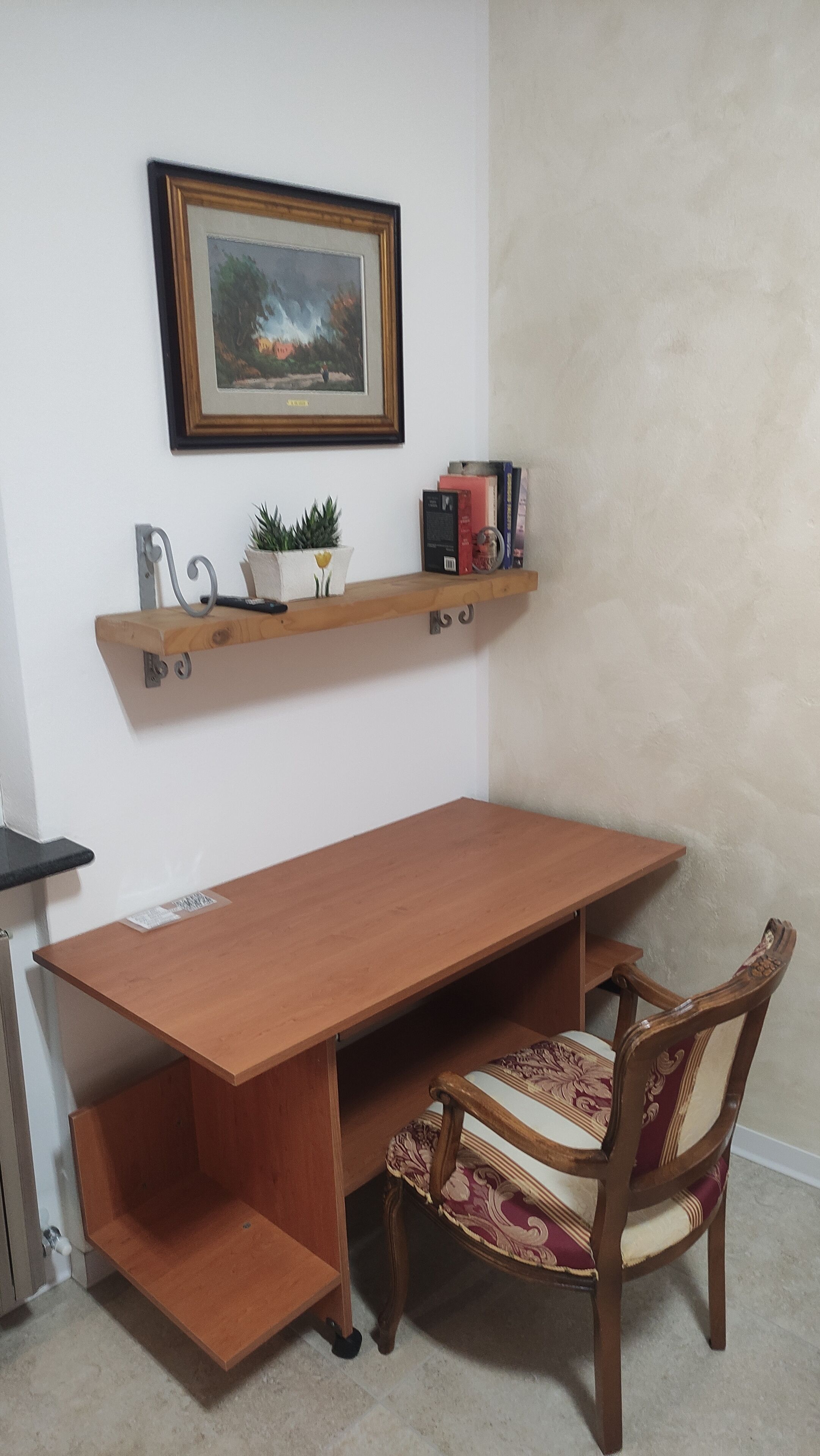 Ein Holztisch mit einem Stuhl, ein Regal mit Büchern und einer Pflanze sowie ein gerahmtes Bild an der Wand.