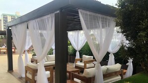 Ein überdachter Sitzbereich im Freien mit gepolsterten Stühlen und Tischen, umgeben von Grünpflanzen und einer Holzpergola.