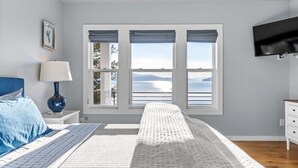 A bedroom with a bed, a nightstand, a lamp, a dresser, a TV, and a window with a view. - Lakefront luxury with dock and views near Gozzer (Harrison)