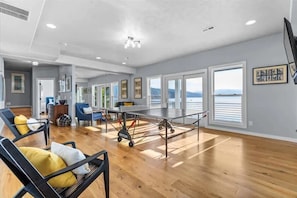 A spacious room with a ping pong table, wooden flooring, and large windows offering a view of the water.