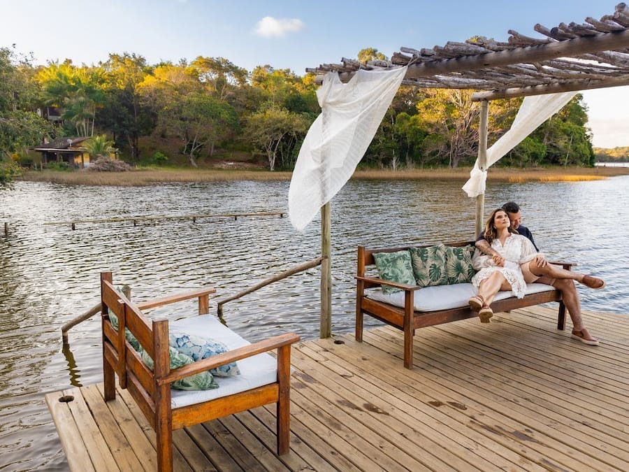 A wooden deck with two wooden chairs and a bench, a canopy, and a view of a lake with trees and a house in the background.